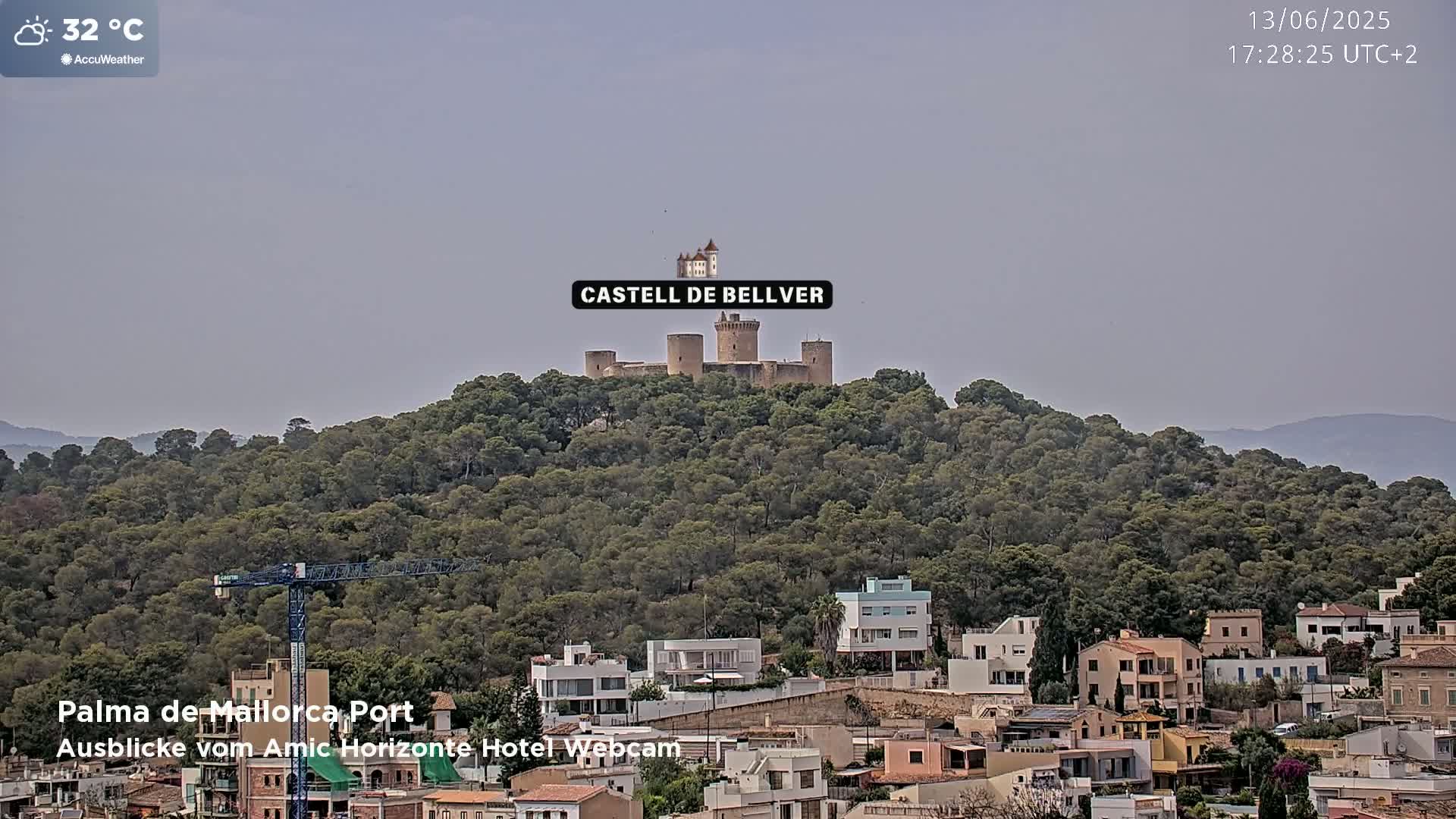 A stone castle sits atop a hill covered in lush green trees, overlooking a town with low-rise buildings under a hazy, light-grey sky, in 32°C weather.
