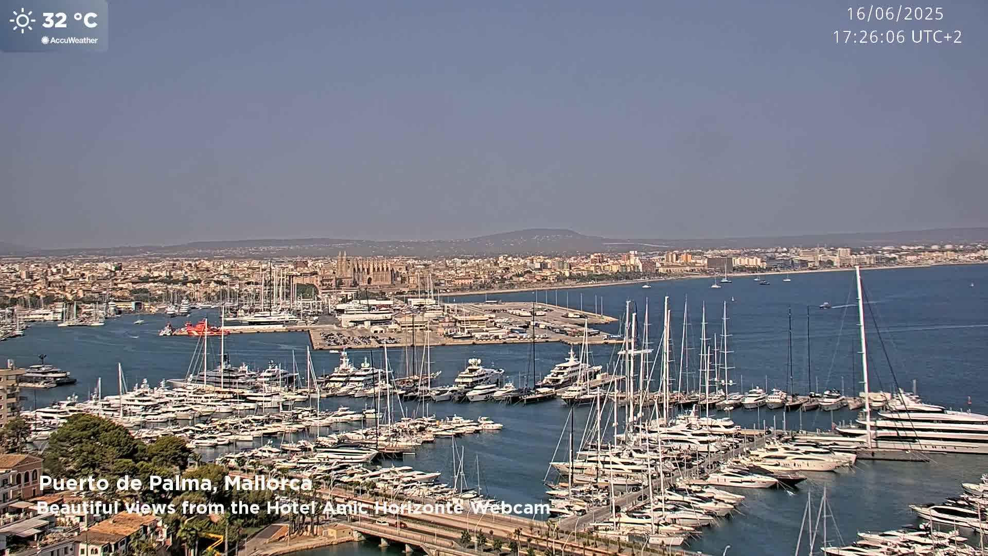 A harbor filled with numerous yachts and sailboats is shown on a sunny day, with a city and a cathedral visible in the background.