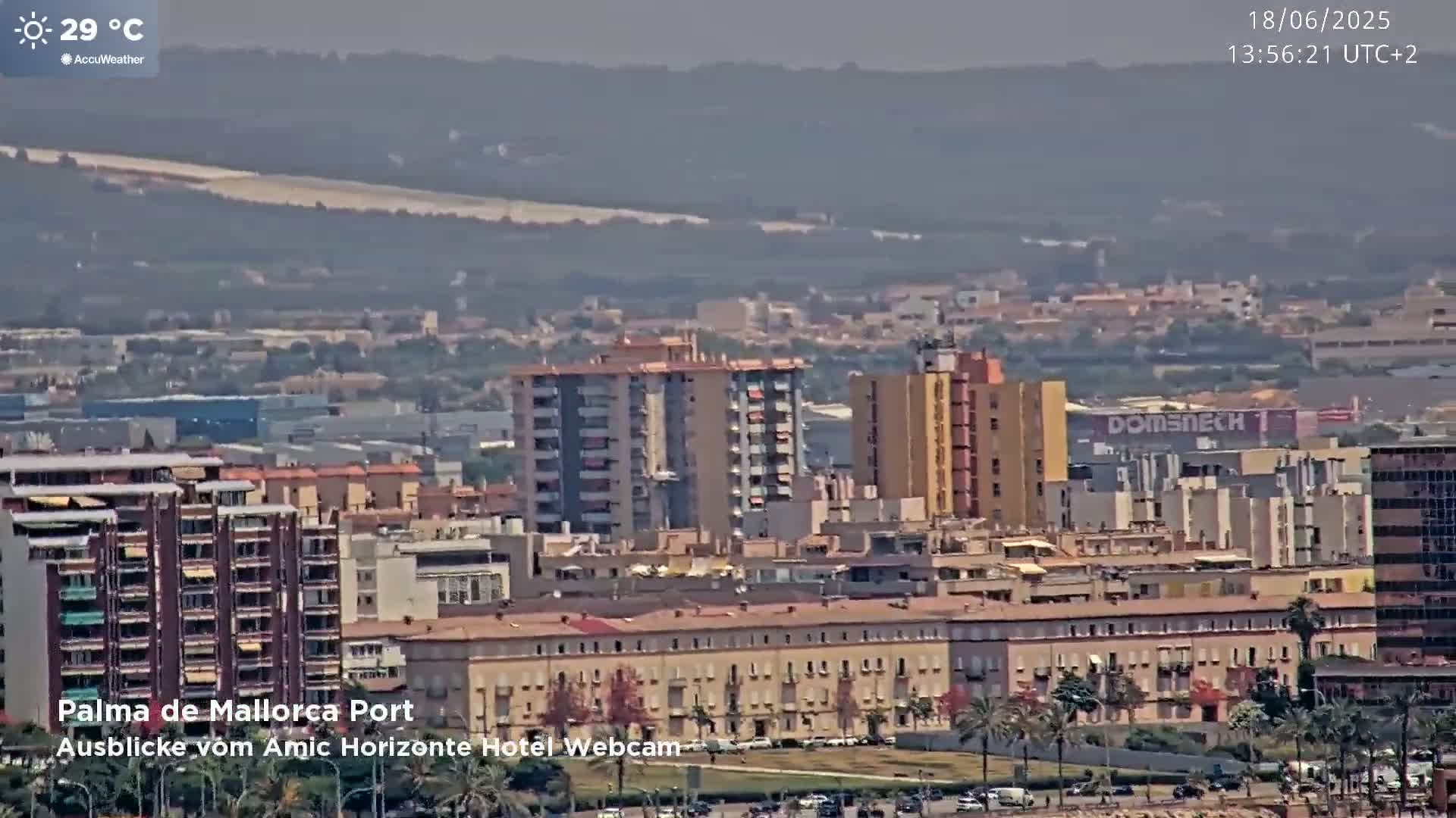 A hazy, sunny view of a city port with numerous buildings, including high-rises and low-rise structures, stretching to distant hills under a clear sky.
