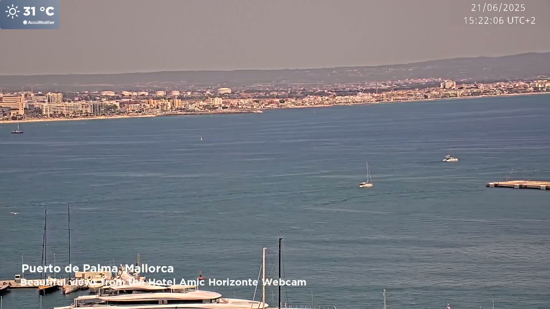 A hazy, sunny day shows a coastal city across a calm body of water with several sailboats and a marina in the foreground.