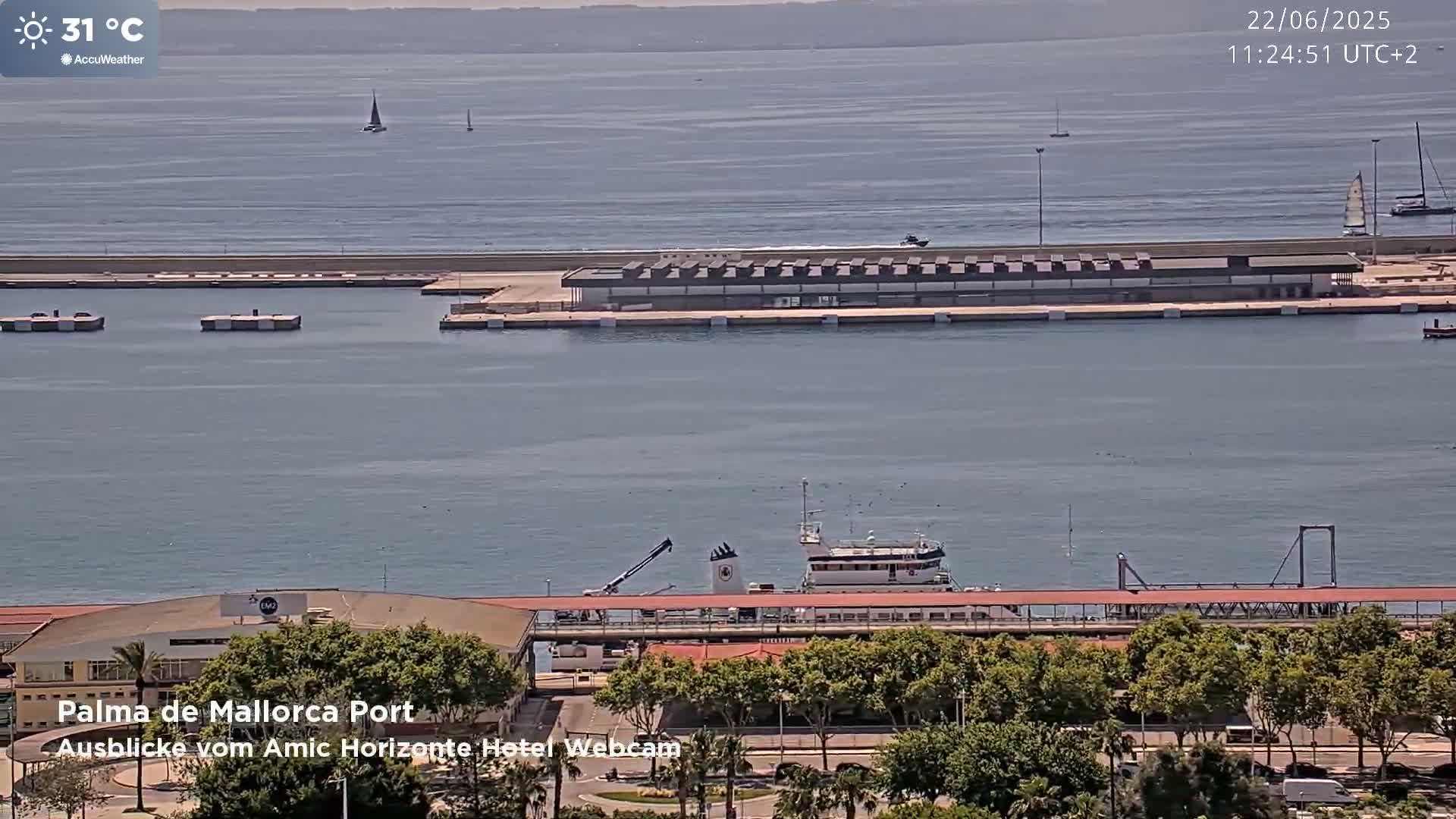 A harbor scene on a sunny day with calm waters shows several boats, a long pier with buildings, and green trees lining a roadway.