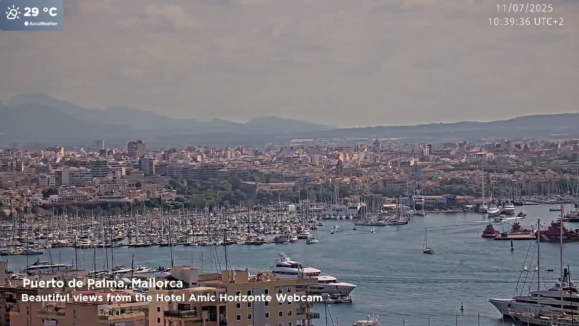 A hazy, 29°C day reveals a cityscape with a large harbor filled with numerous boats and yachts, backed by distant mountains.