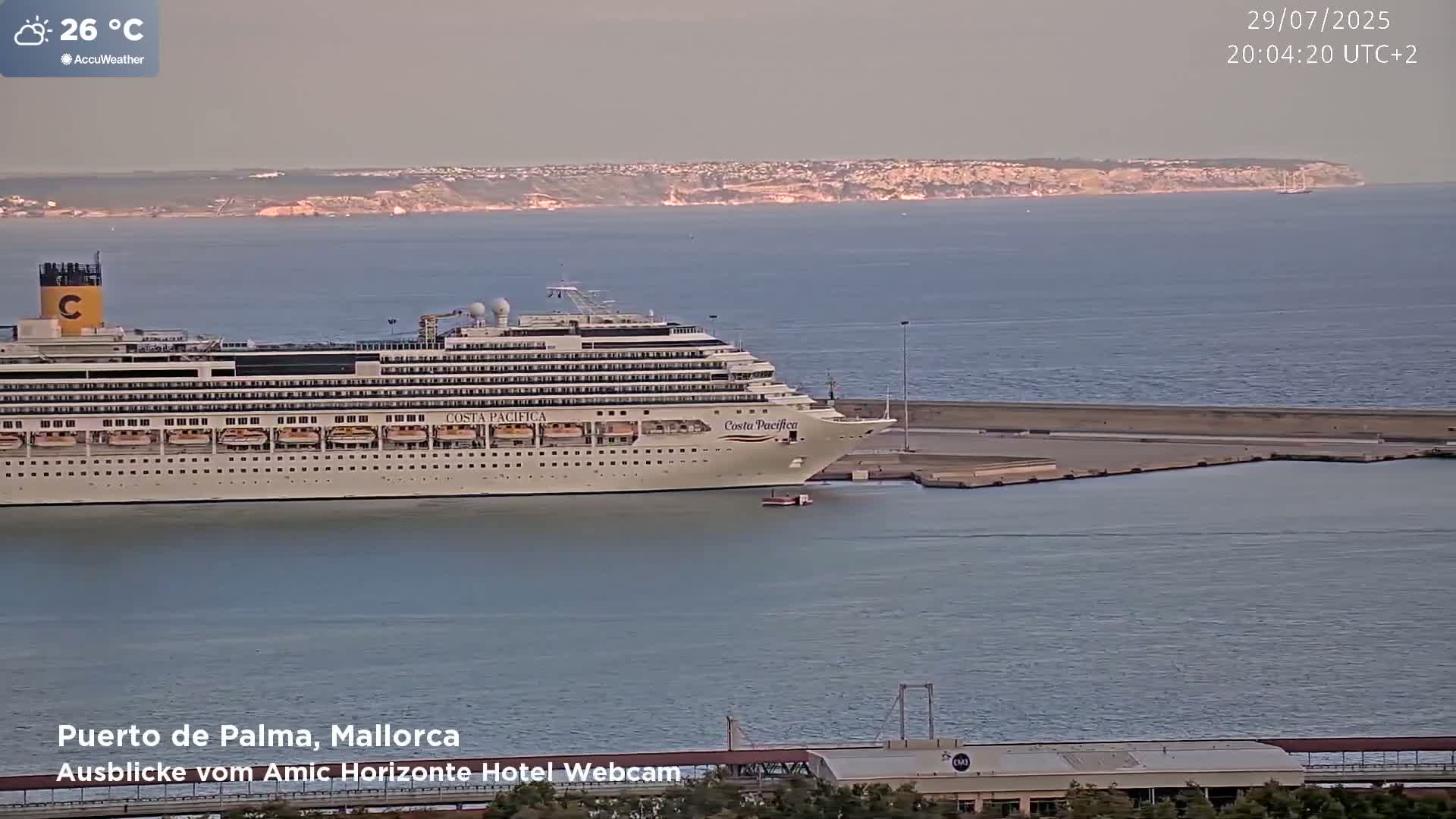 A large cruise ship is docked at a port, with calm water and a distant coastline visible under a hazy sky.