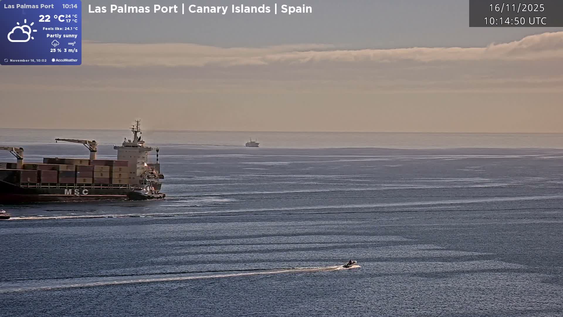 A large container ship, aided by a smaller tugboat, moves across calm waters under a partly sunny sky, while a small speed boat creates a wake in the foreground and another vessel is visible in the distant horizon.