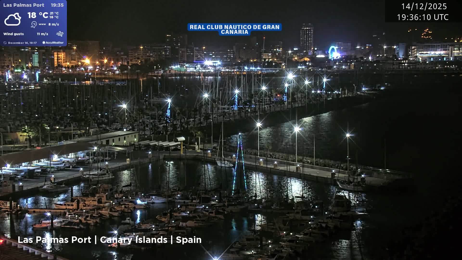 On a cloudy day, a busy multi-lane highway with traffic runs alongside a large marina densely packed with sailboats and yachts, all set against a backdrop of a bustling city and distant mountains.