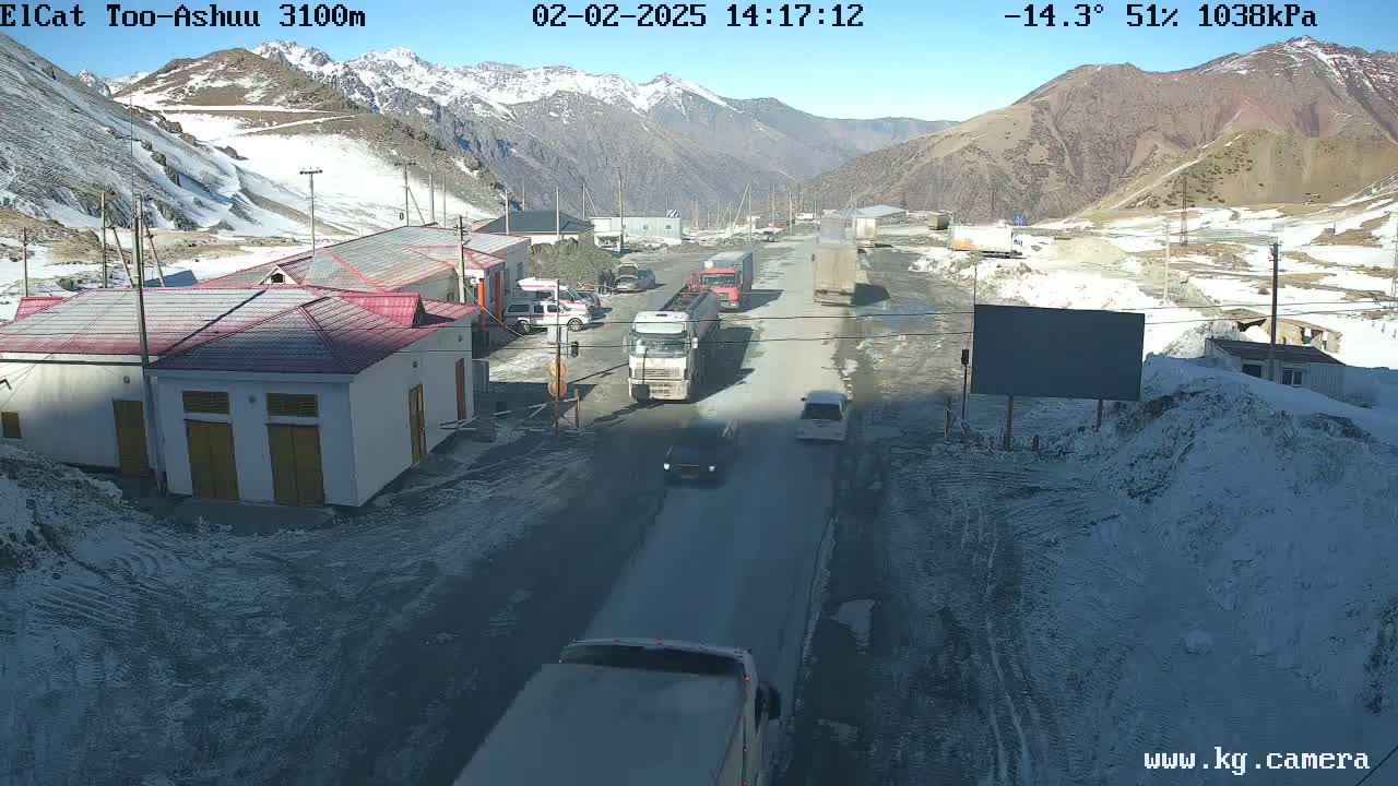 A sunny, cold (-14.3°C) mountain road scene shows several vehicles traveling on a paved road past buildings and snow-covered areas.