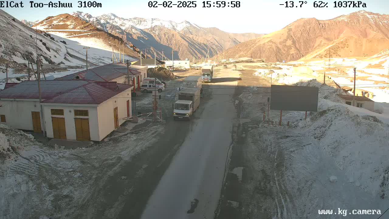 A roadway winds through a snow-covered mountain pass, where several buildings and trucks are visible under a partly sunny sky.