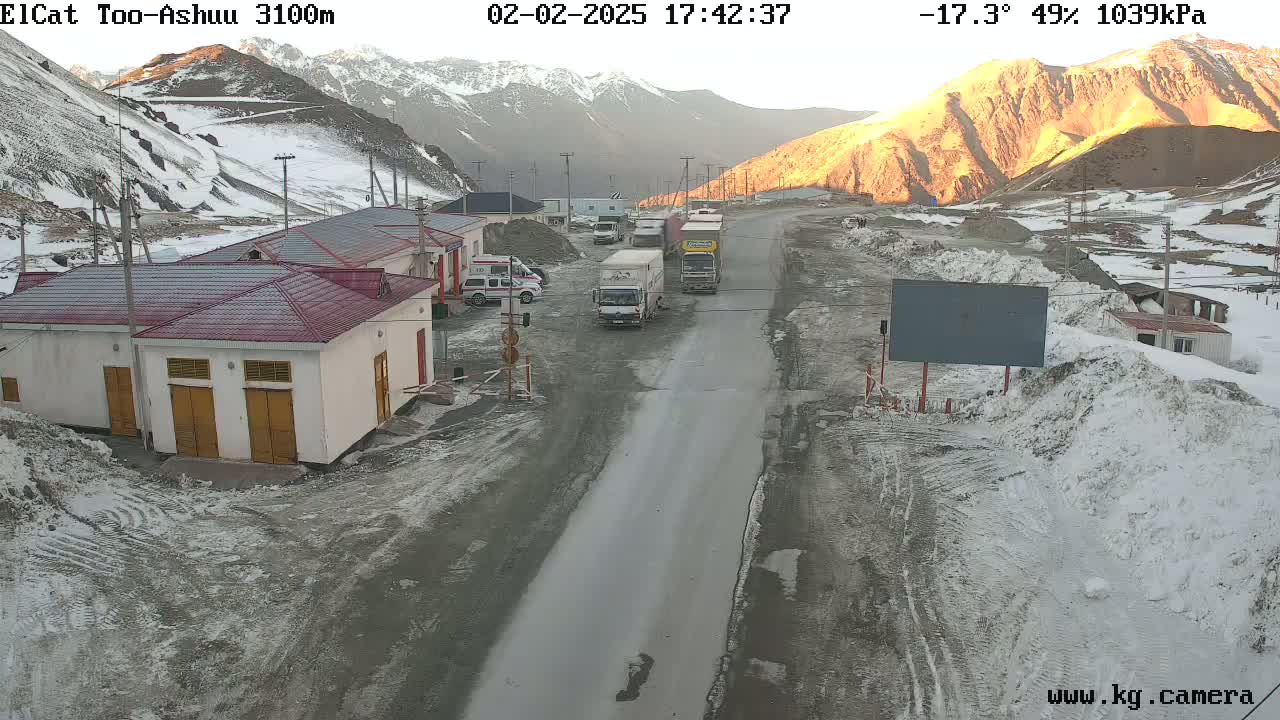A mountain road scene at sunset shows several vehicles parked near buildings in a snowy, mountainous area.