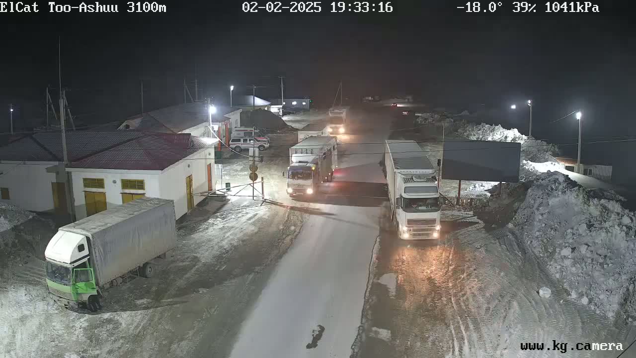 At night in snowy conditions, several trucks are parked or moving slowly along a road near a building complex with snow piles to the side.
