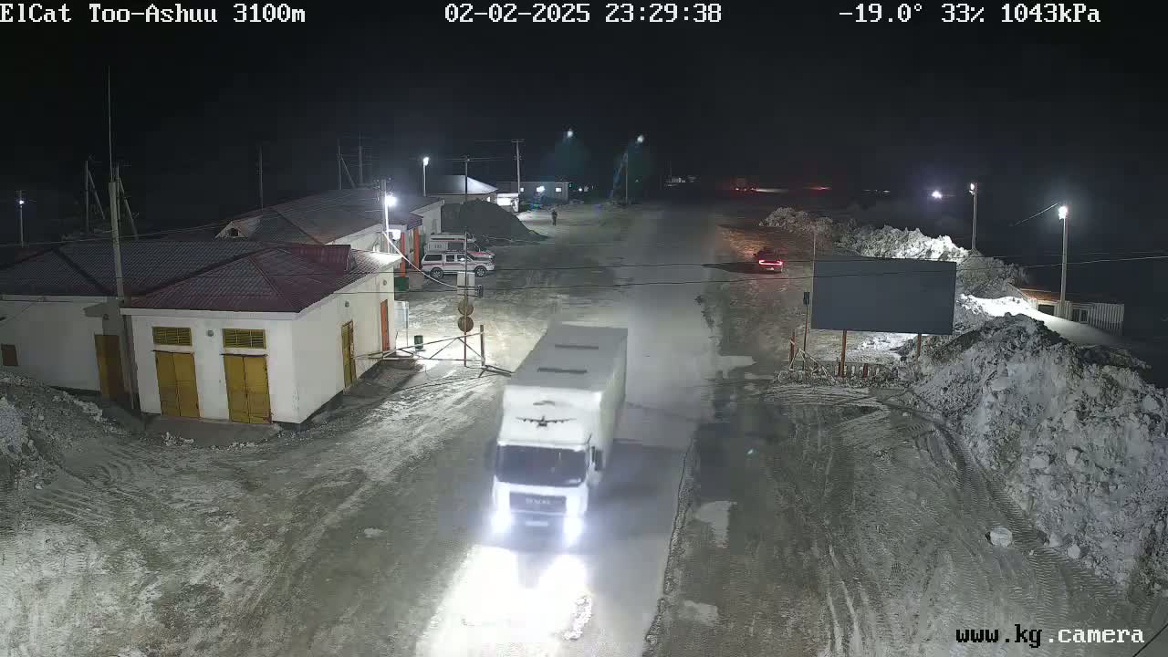 A white truck drives down a snow-covered road at night, past buildings and piles of snow, under a dark, cold sky with a temperature of -19 degrees Celsius.