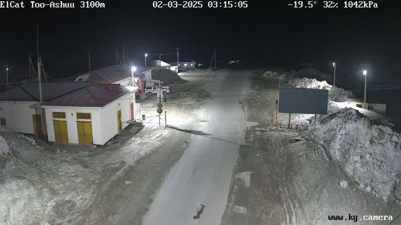 A nighttime view of a snow-covered road in a sparsely populated area with several buildings and vehicles, under artificial light.