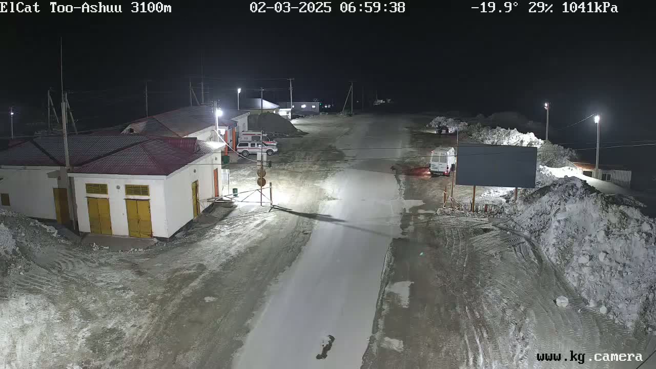 A nighttime, snow-covered street scene shows several buildings and vehicles under artificial light.
