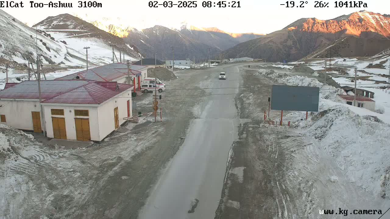 A mountain road scene at sunrise shows a few buildings, some vehicles, and patches of snow and ice on the ground and surrounding mountains in a cold, partly cloudy climate.