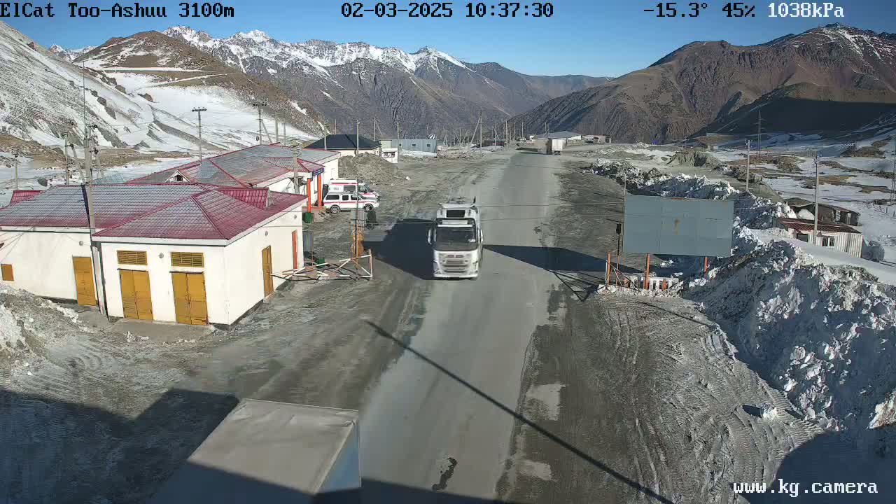 A white truck drives on a road through a mountain pass on a sunny, cold day with snow on the ground and mountains in the background.