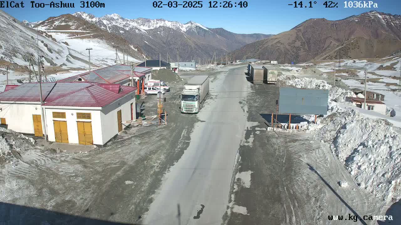 A semi-truck is parked on a road at a mountain pass checkpoint in sunny, cold weather with snow on the ground and surrounding mountains.