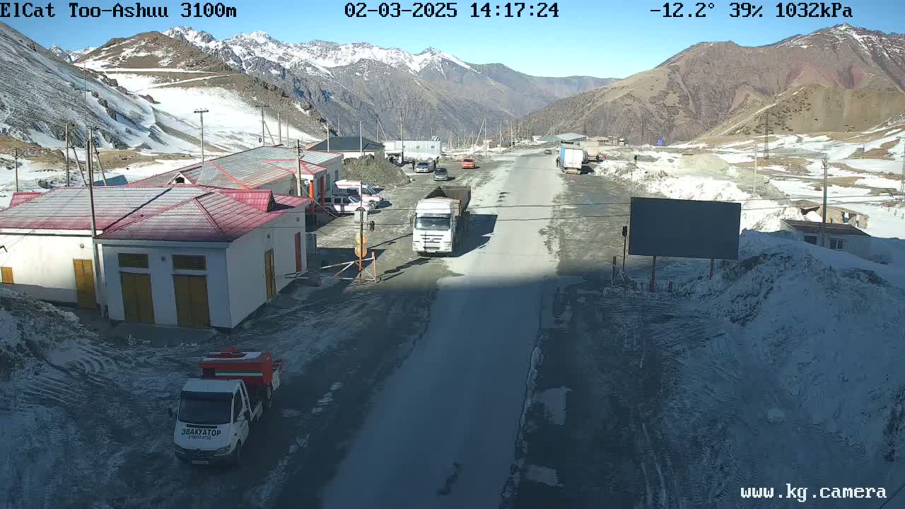 A sunny, cold day at a mountain pass shows several vehicles parked near buildings and a road with a few trucks driving on it, with snow visible on the ground and surrounding mountains.