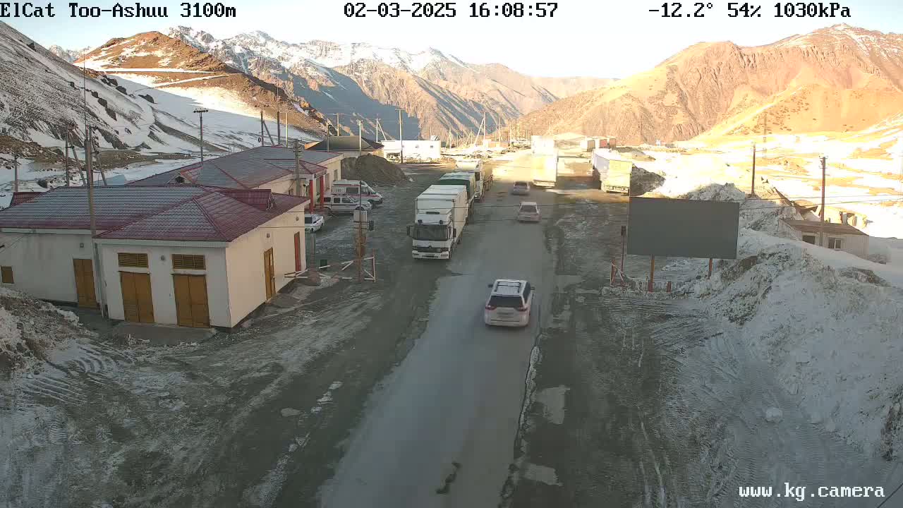 A mountain road scene shows several vehicles, including trucks and cars, near some buildings in a snowy, sunny location.