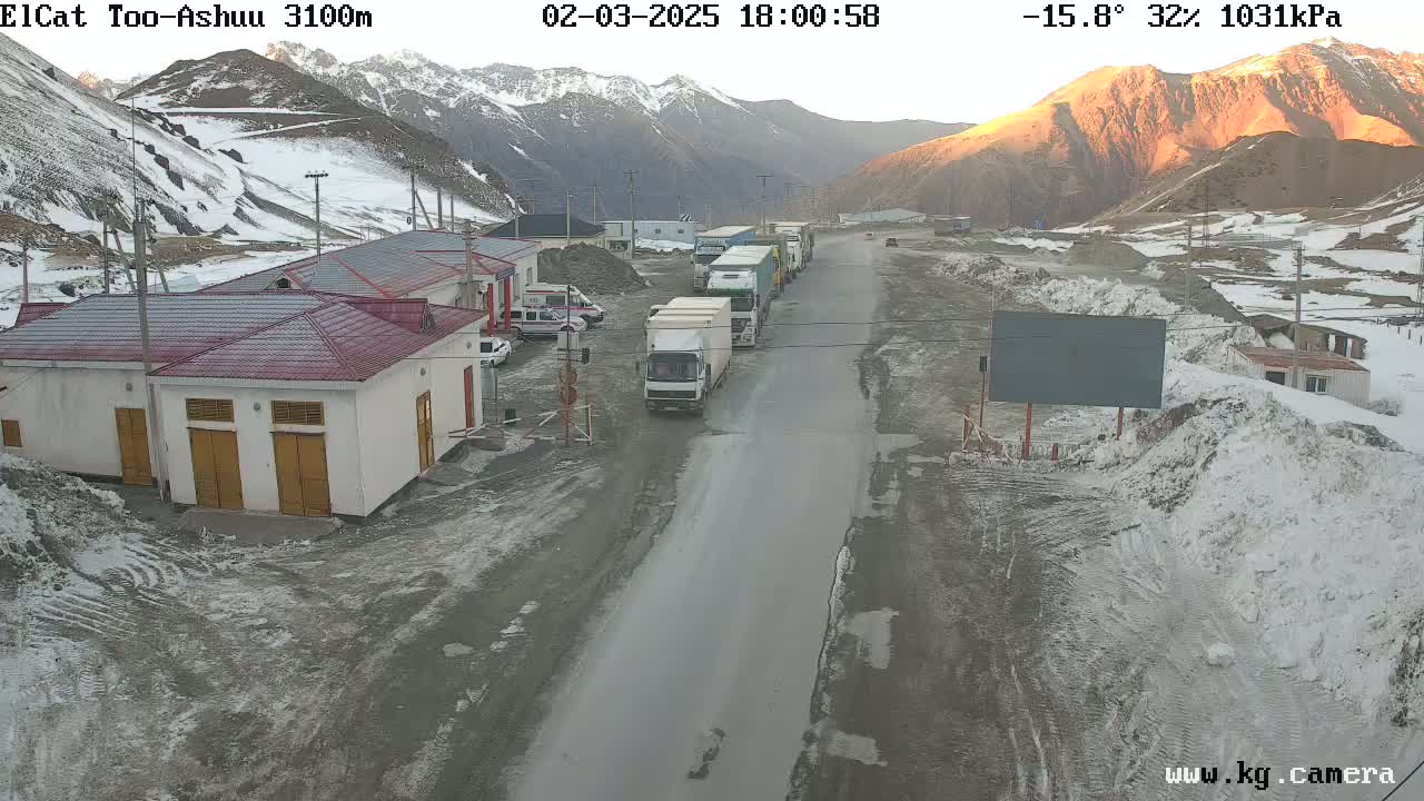 A mountain road with several trucks parked along the side, near some buildings, with snow on the ground and mountains in the background during a cold, partly sunny day.