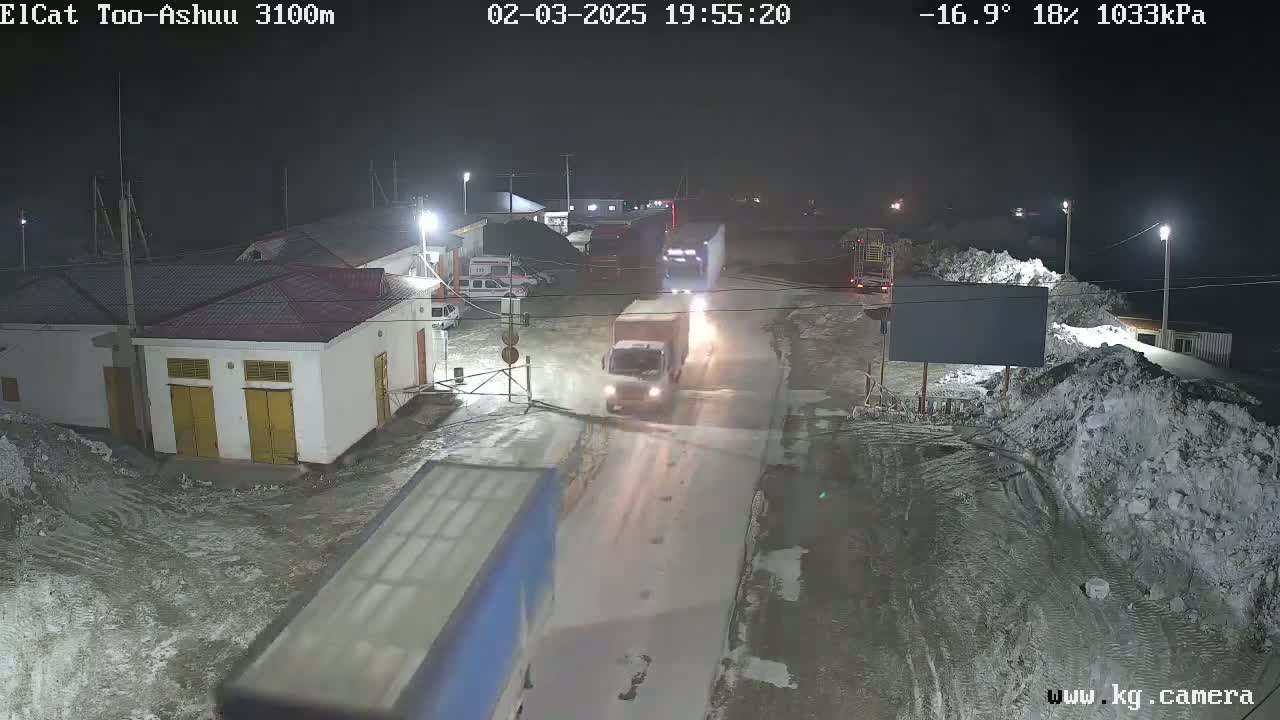 A nighttime, snowy scene shows several trucks and other vehicles on a road near a building complex, under lightly falling snow at -16.9°C.