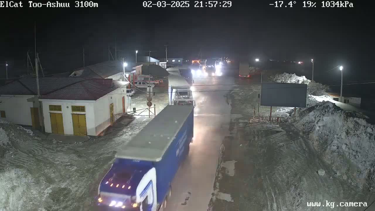 At night, under a dark sky with a temperature of -17.4°C, several trucks are seen driving on a wet, snow-covered road near buildings.
