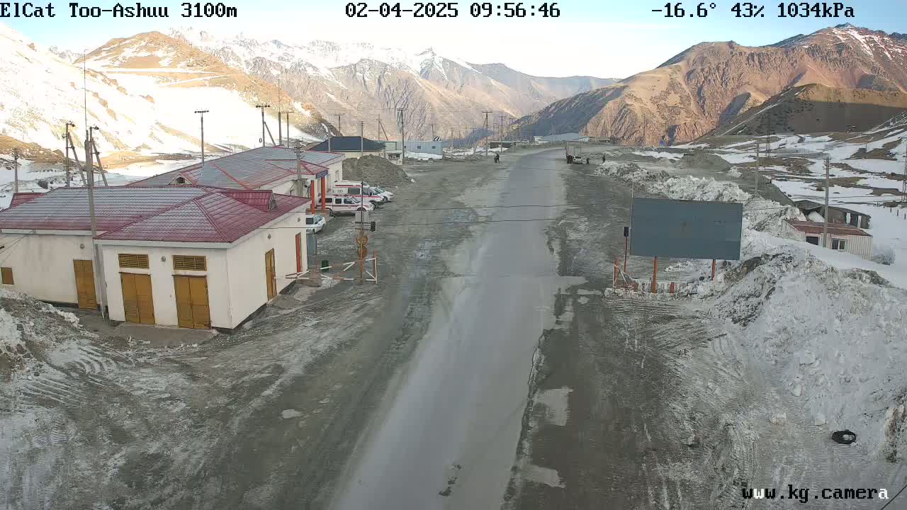 A long, wet road stretches through a mountainous landscape with snow and ice patches, past a few buildings under a partly cloudy sky at a temperature of -16.6°C.
