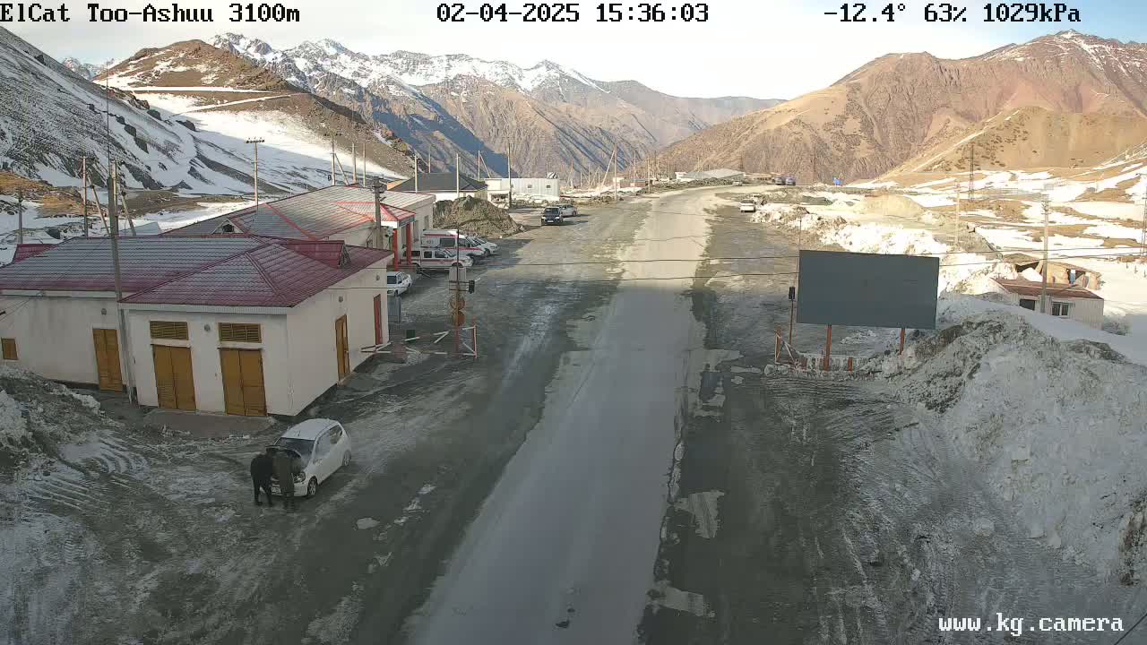 A mountain road scene with snow on the ground and mountains in the background, under a sunny sky.