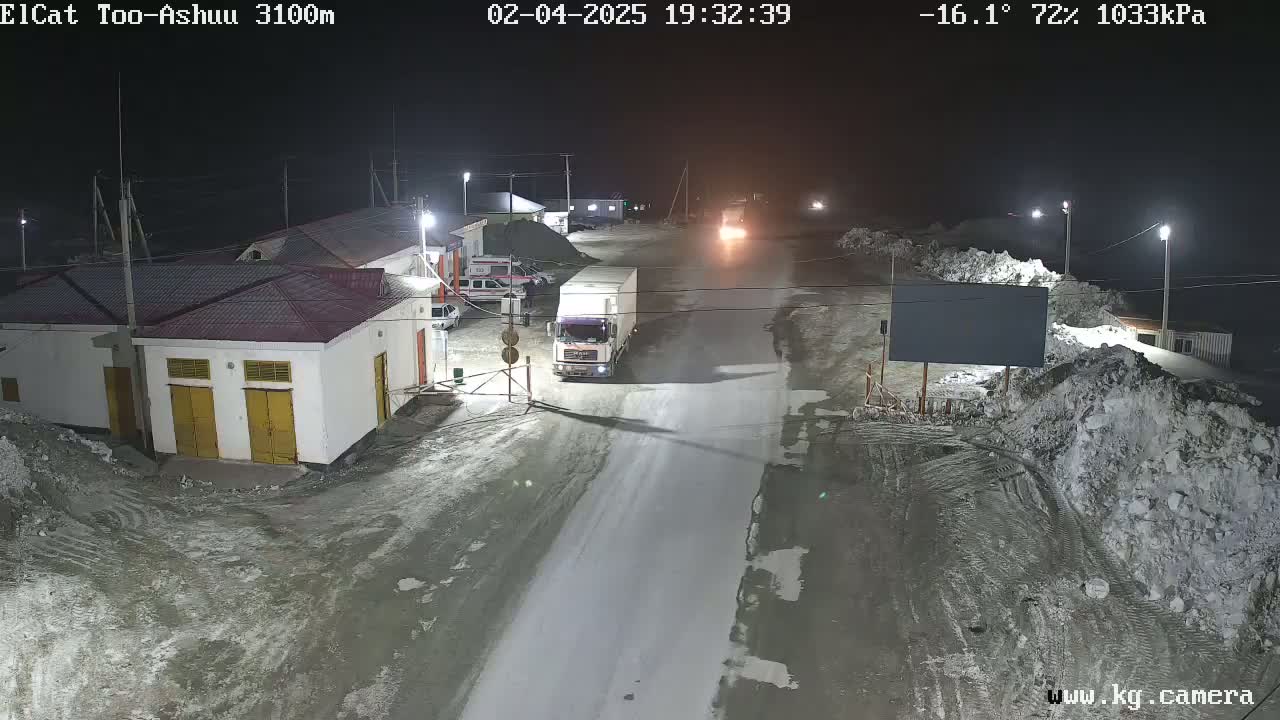 A snow-covered road at night, illuminated by streetlights, shows a truck parked near a building complex with vehicles nearby.