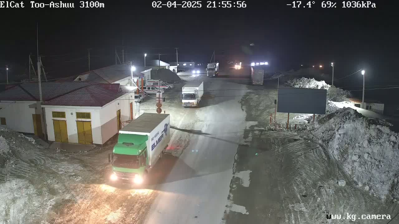 A nighttime, snowy scene shows several trucks parked and driving on a road near buildings, under conditions of -17.4°C, 69% humidity, and 1036 kPa air pressure.