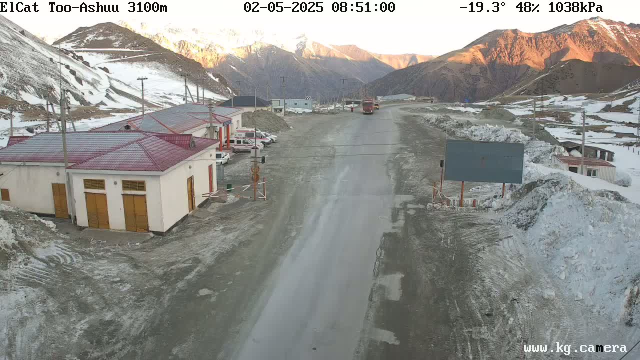 A mountain road scene at sunrise shows several buildings, some vehicles, and snow and ice on the ground and surrounding mountains in cold, partly cloudy conditions.
