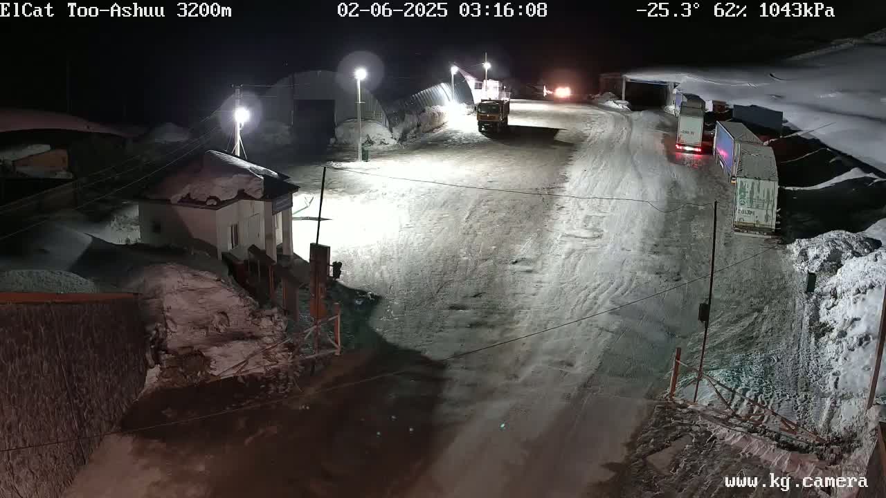 A snow-covered road at night with several trucks parked along the side, near a small building, under a dark sky.