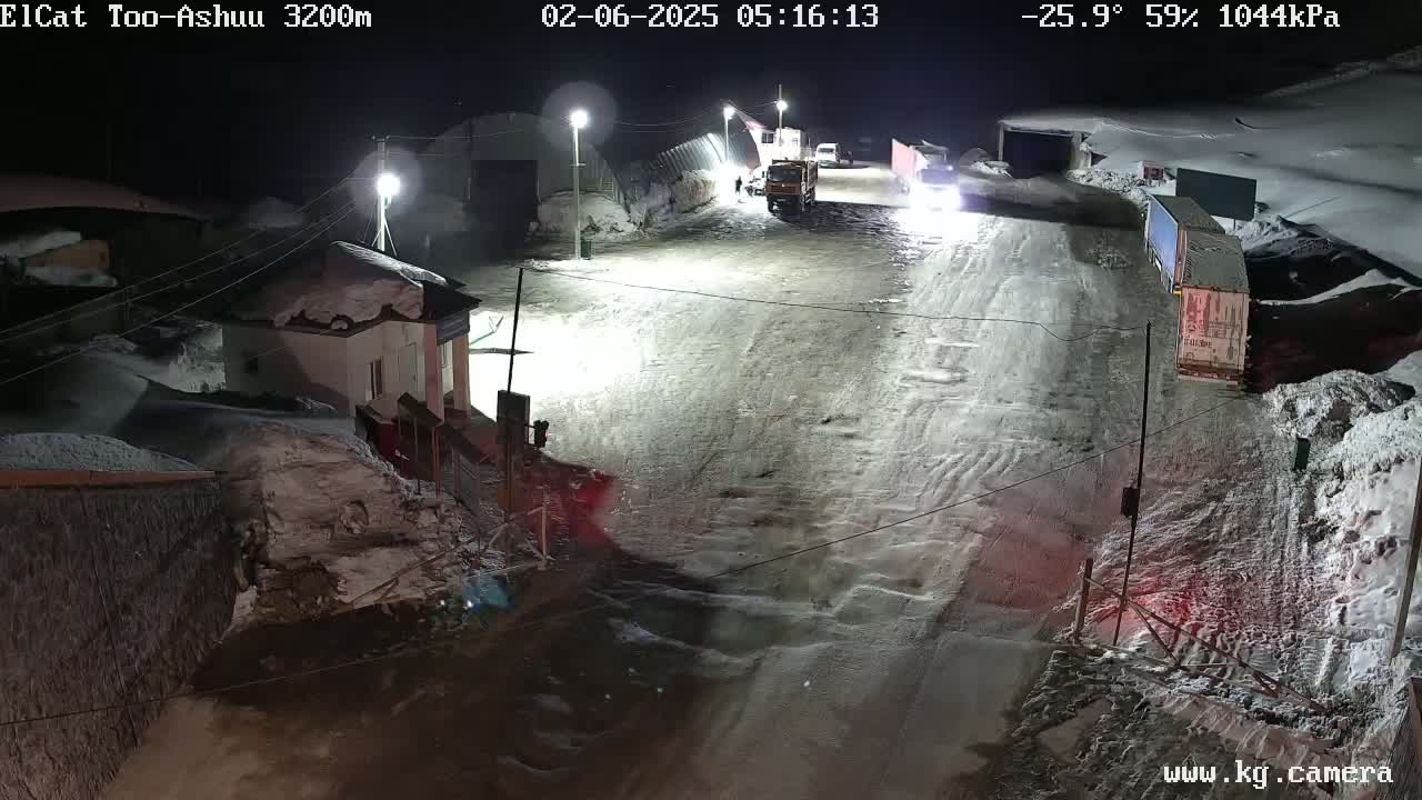 A snow-covered road at night with several vehicles, including trucks and cars, and buildings partially obscured by snow.
