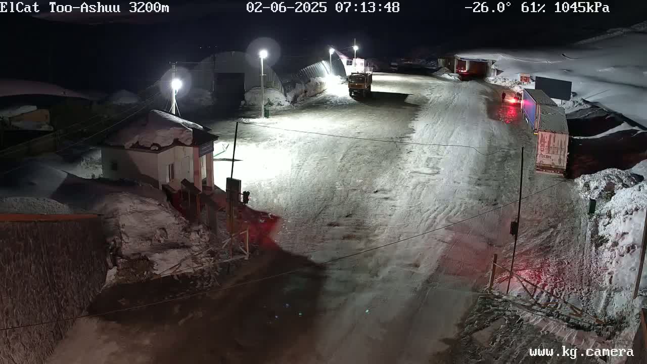 A snow-covered road at night with several trucks and a small building, illuminated by streetlights under a cold, partly cloudy sky.