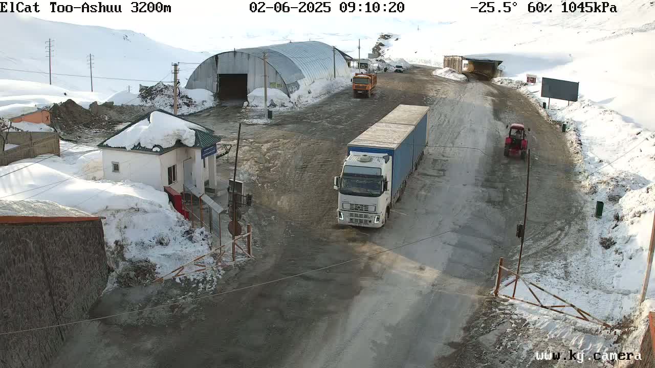 A snowy mountain road scene shows a large semi-truck and a smaller tractor near a checkpoint building with several other vehicles, under a partly sunny sky.