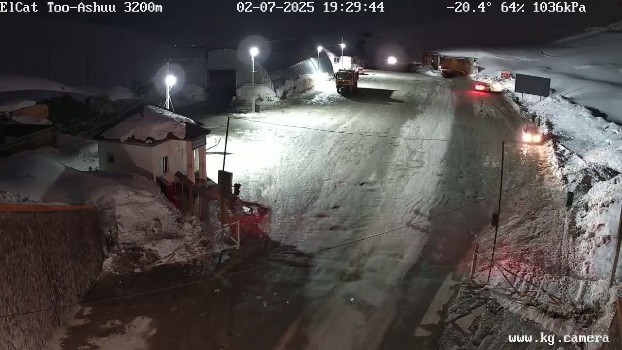 A snow-covered road at night with several vehicles, including a large truck, and buildings in the background under a cold, dark sky.