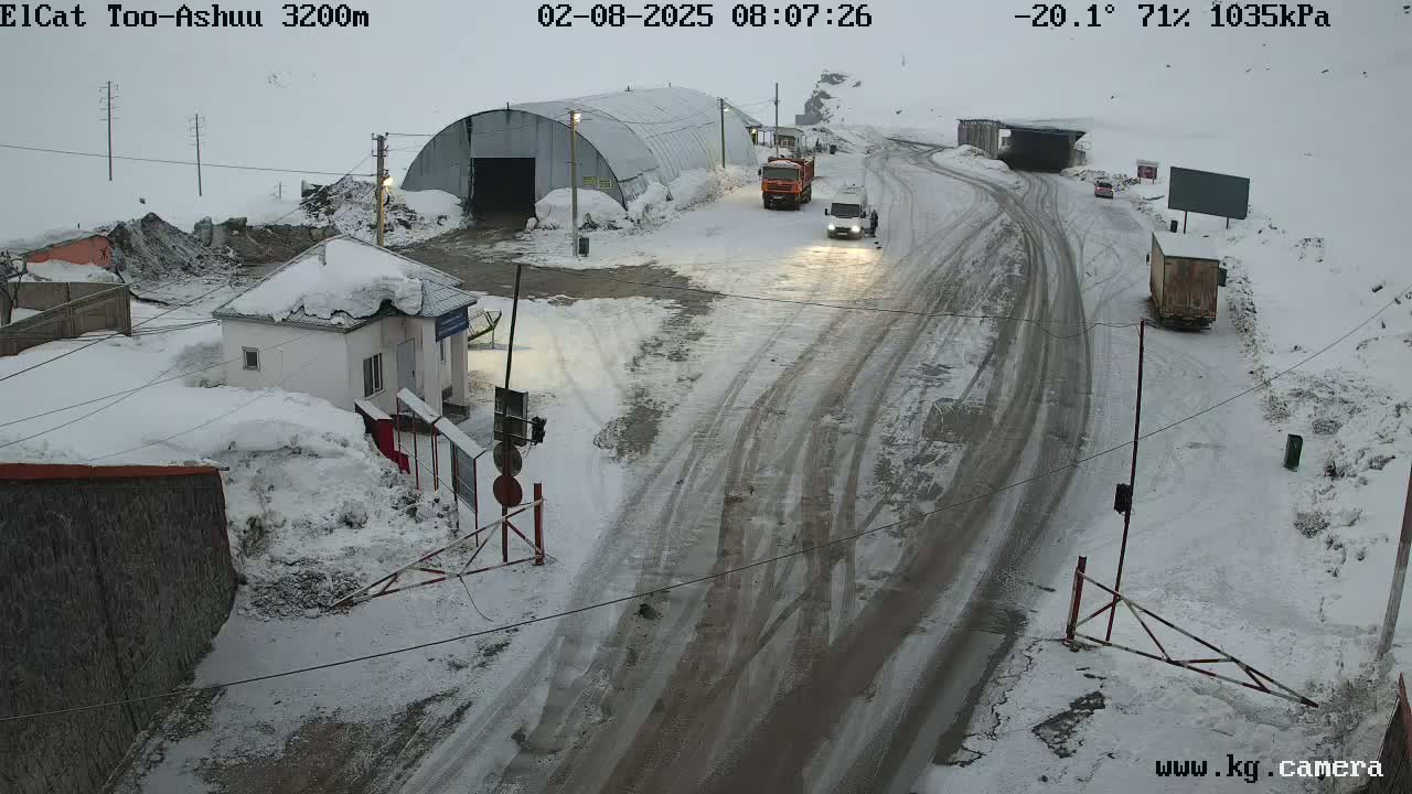 A snow-covered roadway with several vehicles and buildings, including a small structure that appears to be a checkpoint, under overcast skies.