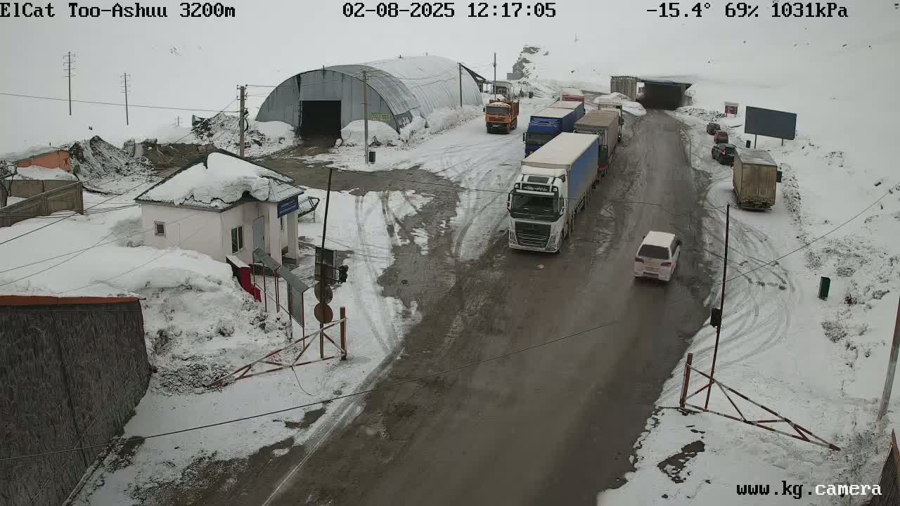 A snowy road with several trucks and cars near a small building and a large metal structure, under a cloudy sky.