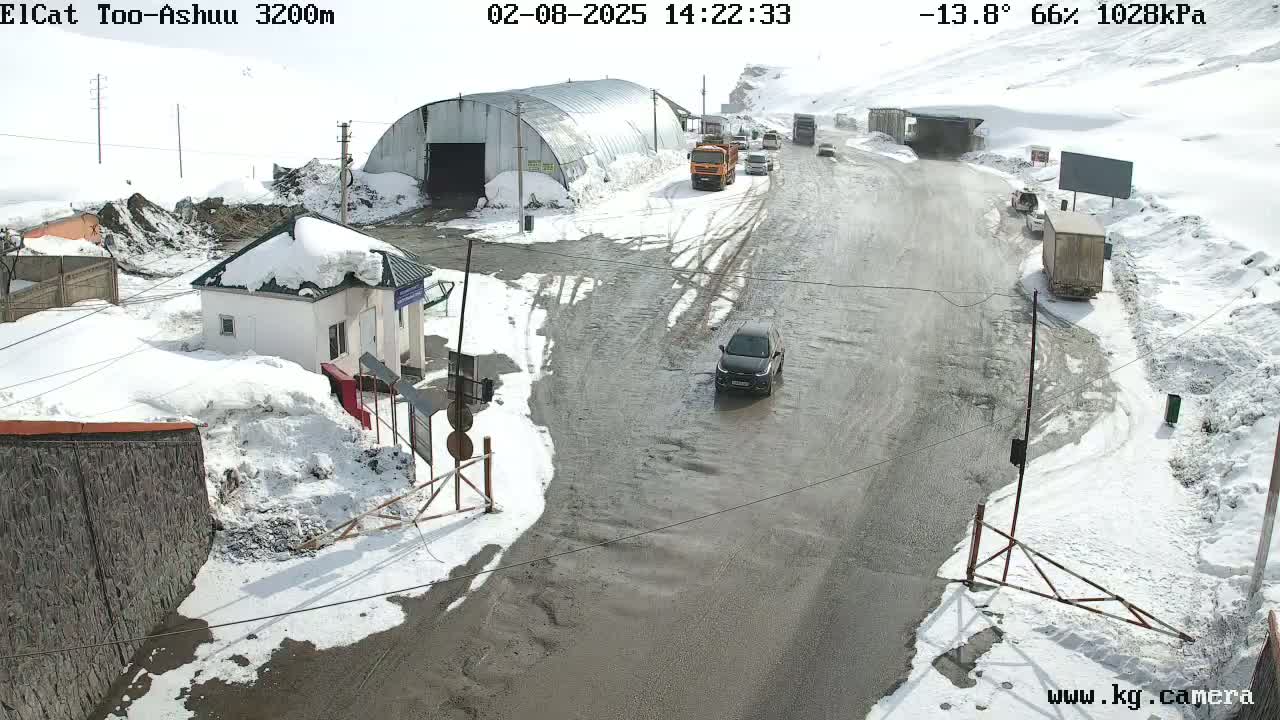 A snow-covered roadway with several vehicles and buildings, under a partly sunny sky.