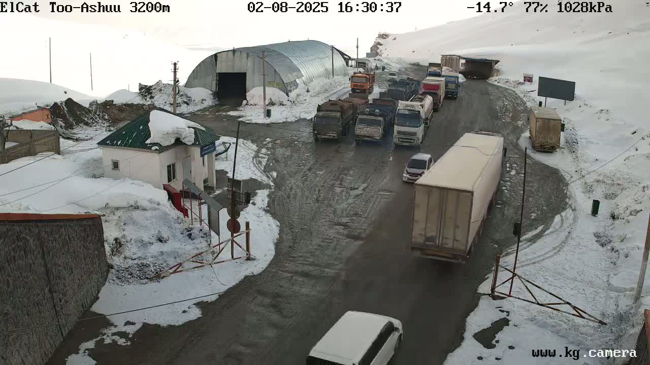 A snowy mountain pass road with several trucks and cars, some stopped at a checkpoint near a small building.