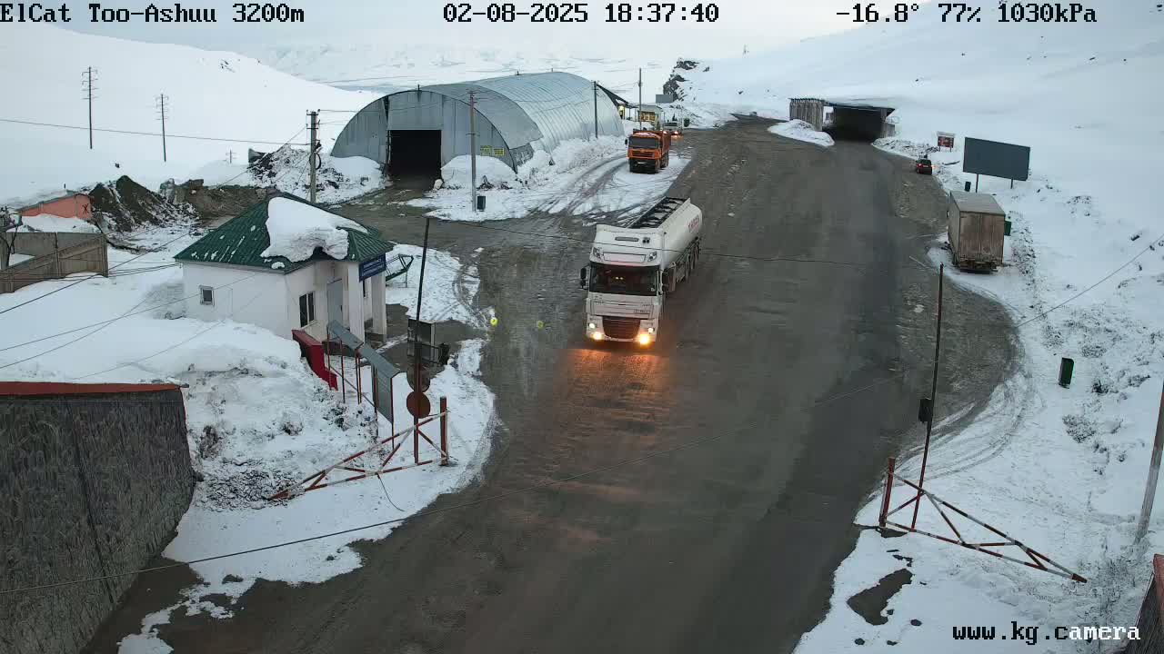 A snow-covered mountain road scene shows several vehicles near a small building and a large metal structure, with a tanker truck in the foreground.