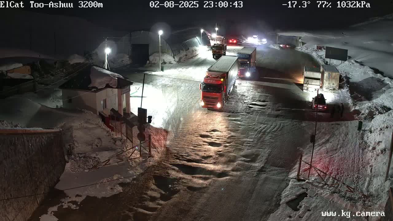 A snowy, nighttime scene shows several trucks and cars stopped on a road near buildings, with a temperature of -17.3°C and 77% humidity.