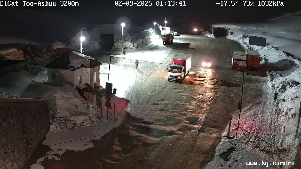 A nighttime scene of a snow-covered roadway with several trucks and a small building, under a cold, partially cloudy sky.