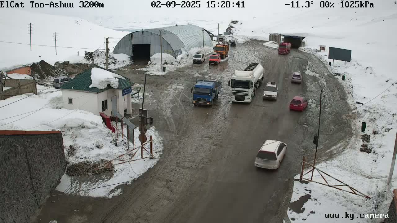 A muddy road with several vehicles, including trucks and cars, is seen near a small building and a large metal structure in a snowy, overcast landscape.