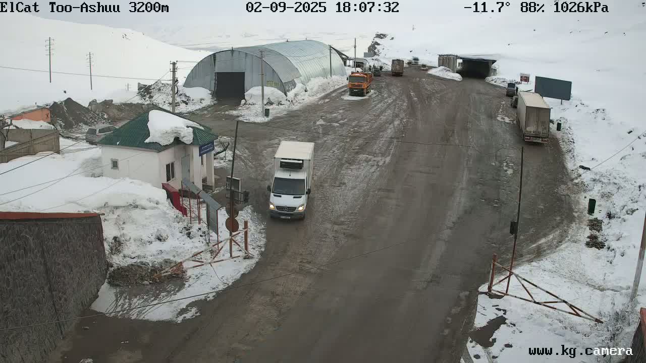 A snowy, overcast scene shows a roadway with several trucks and a small building, possibly a border crossing, with snow covering the ground and some buildings.