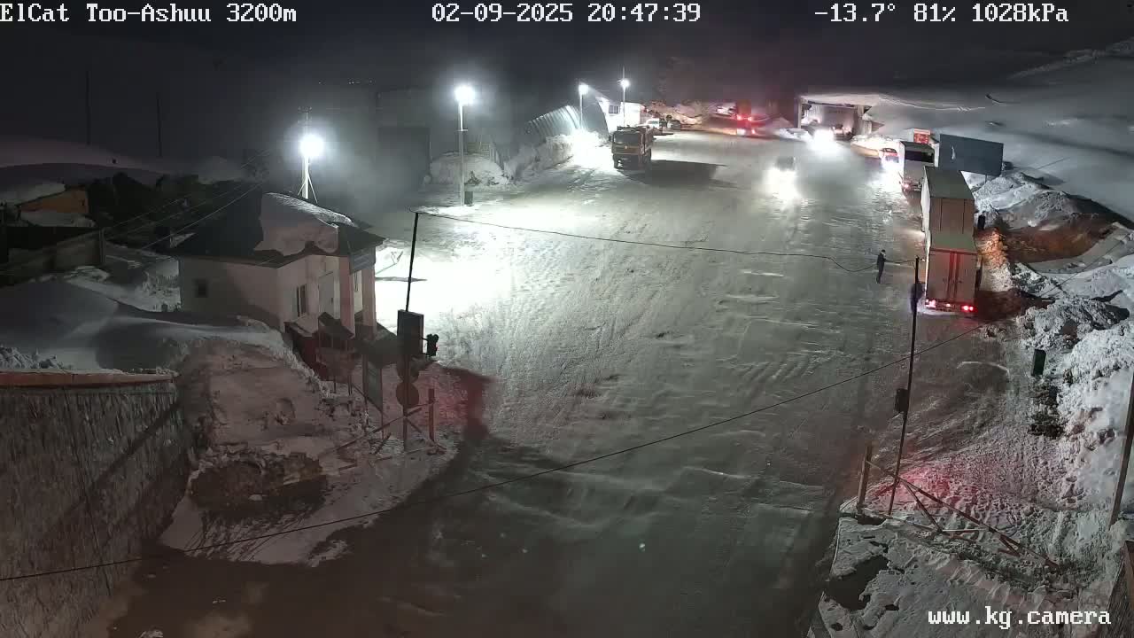 A nighttime, snowy scene shows several vehicles parked and stopped at a crossroads near a building, under partly cloudy skies and a temperature of -13.7°C.