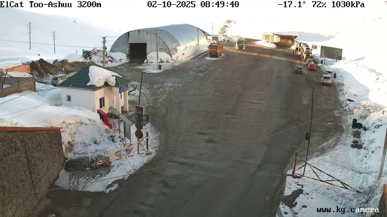 A snow-covered roadway with several vehicles, including trucks and cars, near a small building and a large metal structure, in -17.1°C weather with 72% humidity.