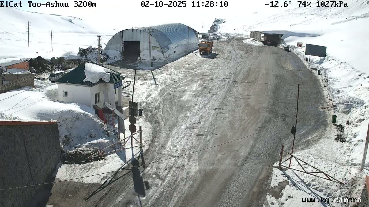 A snow-covered mountain road winds past a small building and a large metal structure, with a few vehicles visible under a sunny sky.