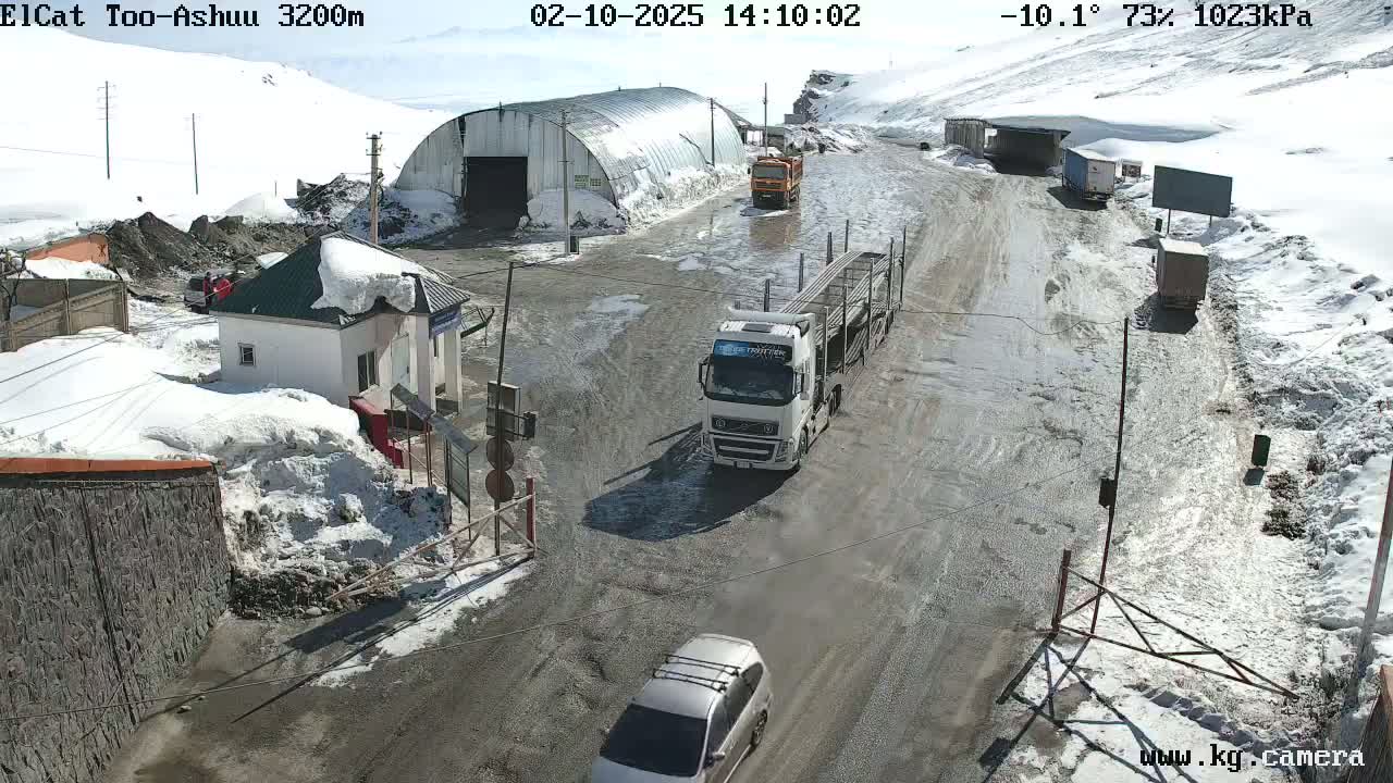 A snow-covered mountain road with several vehicles, including a large truck and a car, near a small building and a larger metal structure, under a sunny sky.