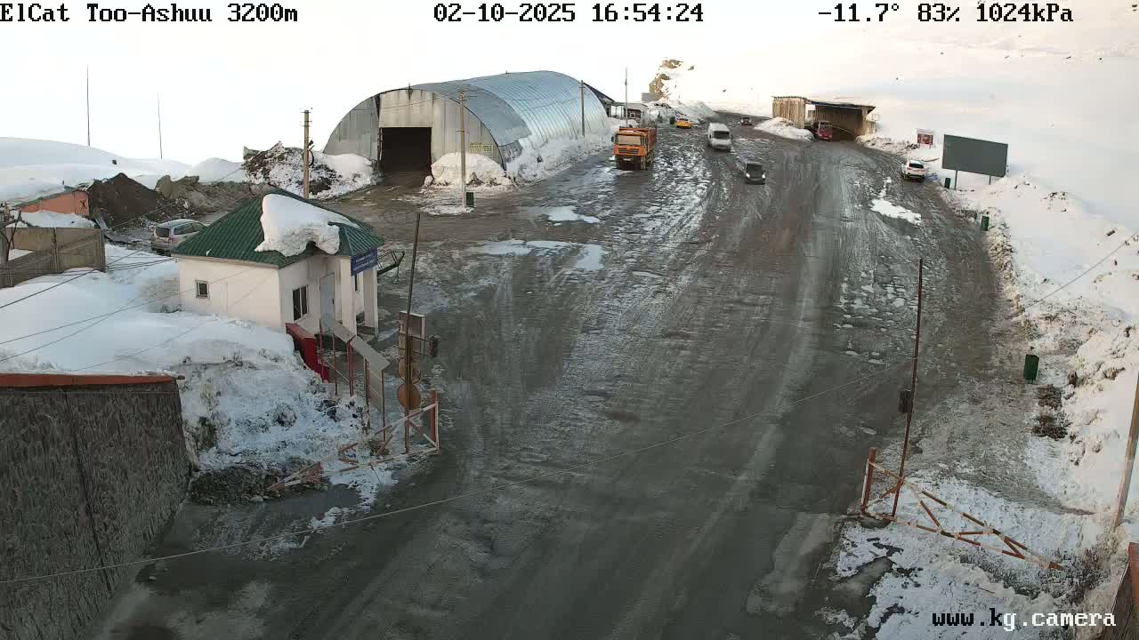 A snow-covered mountain road with several vehicles, a small building, and a large metal structure, under cloudy skies at -11.7°C.