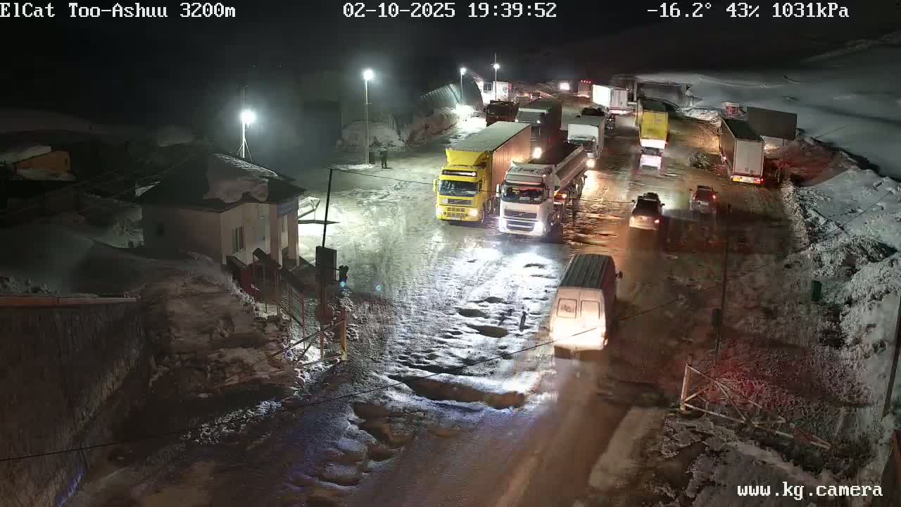 A nighttime scene shows a line of vehicles, including several large trucks, stopped on a snow-covered and partially icy road with potholes, under a partly cloudy sky at -16.2°C.