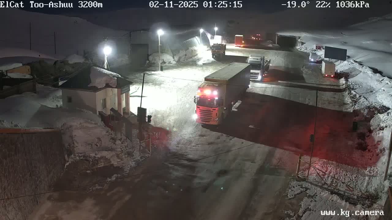 Several large trucks are parked in a snowy nighttime roadway near buildings, with snow piled high along the sides.